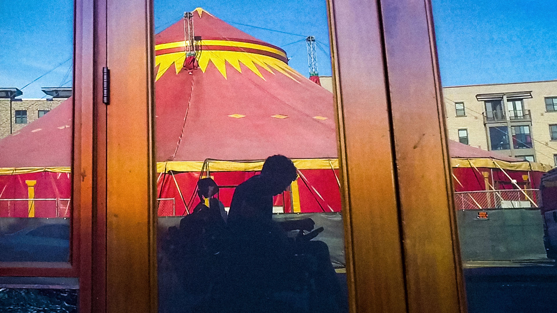 In a reflection of an unmarked storefront is a grayish silhouette of a man using an electric wheelchair. Behind the man is a spectacular red and yellow circus tent. Image credit: Reid Davenport