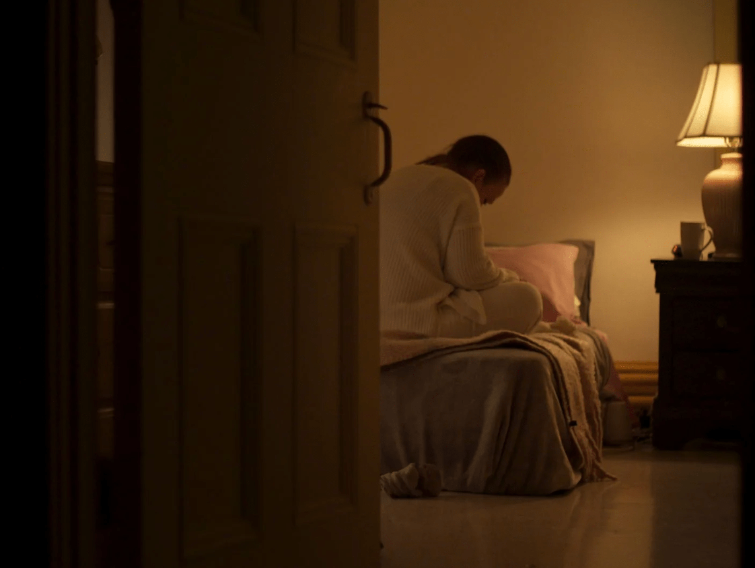 A woman sits alone on a bed in a dimly lit room, gazing downward, with a lamp casting a warm glow beside her.