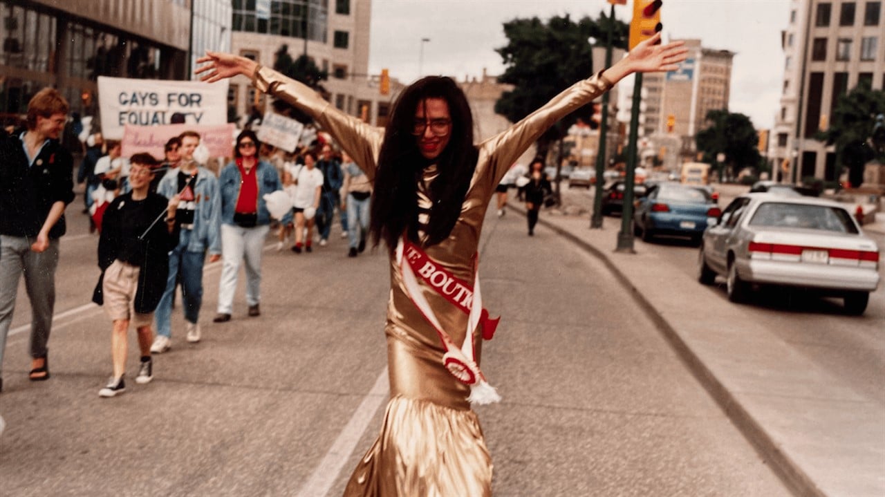 Still from the documentary Parade: Queer Acts of Love & Resistance showing an drag performer in a gold gown leading an LGBTQ+ rights parade with raised arms.