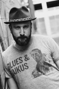 Portrait of filmmaker Noam Gonick wearing a hat and a "Blues & Anxious" T-shirt, shot in black and white.