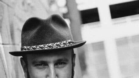 Portrait of filmmaker Noam Gonick wearing a hat and a "Blues & Anxious" T-shirt, shot in black and white.