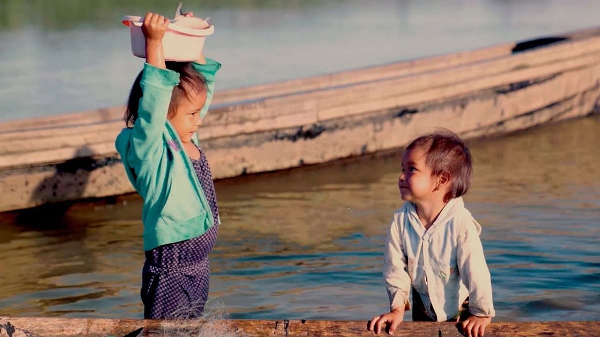 Two children standing in shallow river water beside a wooden boat.
