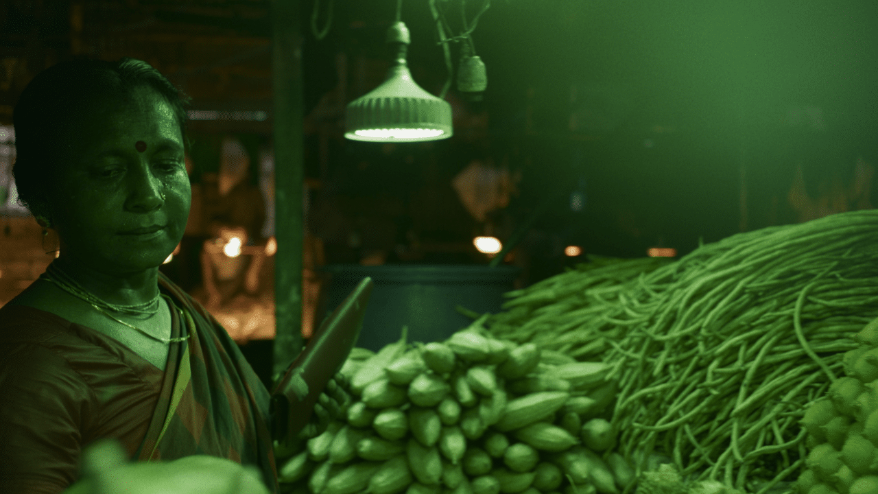 A person in low green light beside piles of produce under a hanging lamp.